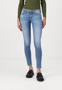 Γυναίκα που φοράει ανοιχτό μπλε στενά τζιν, μαύρες λοφίες και ένα ελαιόχρωμο ριμπ κορυφή με μακριά μανίκια, που στέκεται μπροστά σε λευκό φόντο.