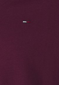 Burgundská mikina vyrobená z měkké tkaniny. Má malé vyšívané logo v černé, bílé a červené barvě na hrudi. Design s výstřihem do V.