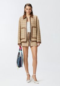 Chaqueta de cuadros beige con detalles en cuero marrón, combinada con una blusa blanca de botones y una mini falda de cuadros a juego. Lleva una bolsa multicolor.