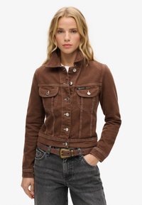 Veste en denim marron au design ajusté, deux poches poitrine, boutons en métal et couture blanche contrastante. Portée sur un haut blanc et un jean gris.