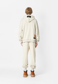 Sudadera beige con capucha grande y hombros caídos; presenta un parche de dibujos animados y una etiqueta en la parte posterior. Joggers beige a juego y botas marrones.