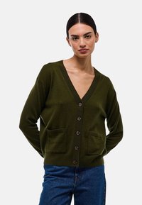 Cardigan vert olive avec un col en V, fermeture à boutons et deux poches avant. Texture douce et coupe décontractée. Associé à un jean bleu.