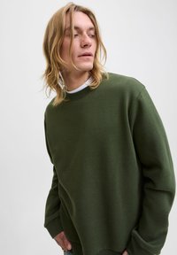 Ung person med långt blont hår, iklädd en oversized mörkgrön sweatshirt, ser åt sidan mot en enkel ljus bakgrund.