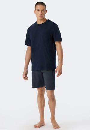 Navy-blaue Baumwoll-T-Shirt mit Brusttasche und gemusterten navyblauen Shorts mit weißen Punkten. Das Modell steht barfuß.