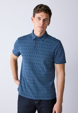 Noor mees lühikeste pruuni juustega, kes kannab tekstuurse sinise lühikeste varrukatega polo särki ja tumedaid pükse, seisab tavalise hele tausta ees.