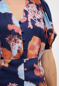 Blumenmusterkleid in Marineblau, mit lebhaften orangen und blauen Farbtönen, puffärmelig und mit stoffüberzogenen Knöpfen auf der Vorderseite.
