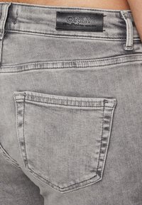 Grå denimjeans med en baklomme, med en svart "ONLY"-label over. Stoffet har en glatt, noe slitt tekstur.