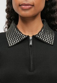 Melnā ribota blūze ar rāvējslēdzēja priekšu un punktētām apkaklēm. Apkakle ir rotāta ar metāliskiem kniedēm noteiktā rakstā. Gluda auduma tekstūra.