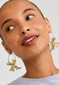 Orecchini a cerchio in tono oro con un design floreale, con petali texturizzati e una finitura liscia, su uno sfondo neutro.