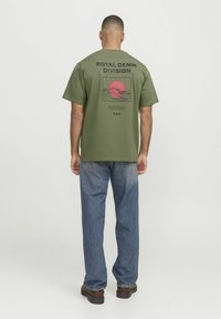 Camiseta de manga corta color verde oliva con texto negro "ROYAL DENIM DIVISION" y un gráfico de sol rojo. Combinada con pantalones de mezclilla azules y zapatos marrones.