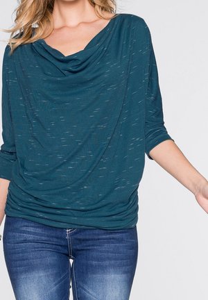 Donna che indossa una maglia color teal con scollo drappeggiato, maniche a tre quarti e jeans aderenti blu su uno sfondo chiaro e uniforme.