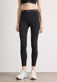 Čierne legíny s vysokým pásom, vyznačujúce sa bielym logom Calvin Klein, vyrobené z hladkého, elastického materiálu, kombinované s svetlými teniskami.