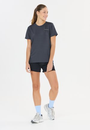 Ung kvinde står og smiler, iført en mørkegrå sports-T-shirt, sorte shorts, lyseblå sokker og hvide løbesko.
