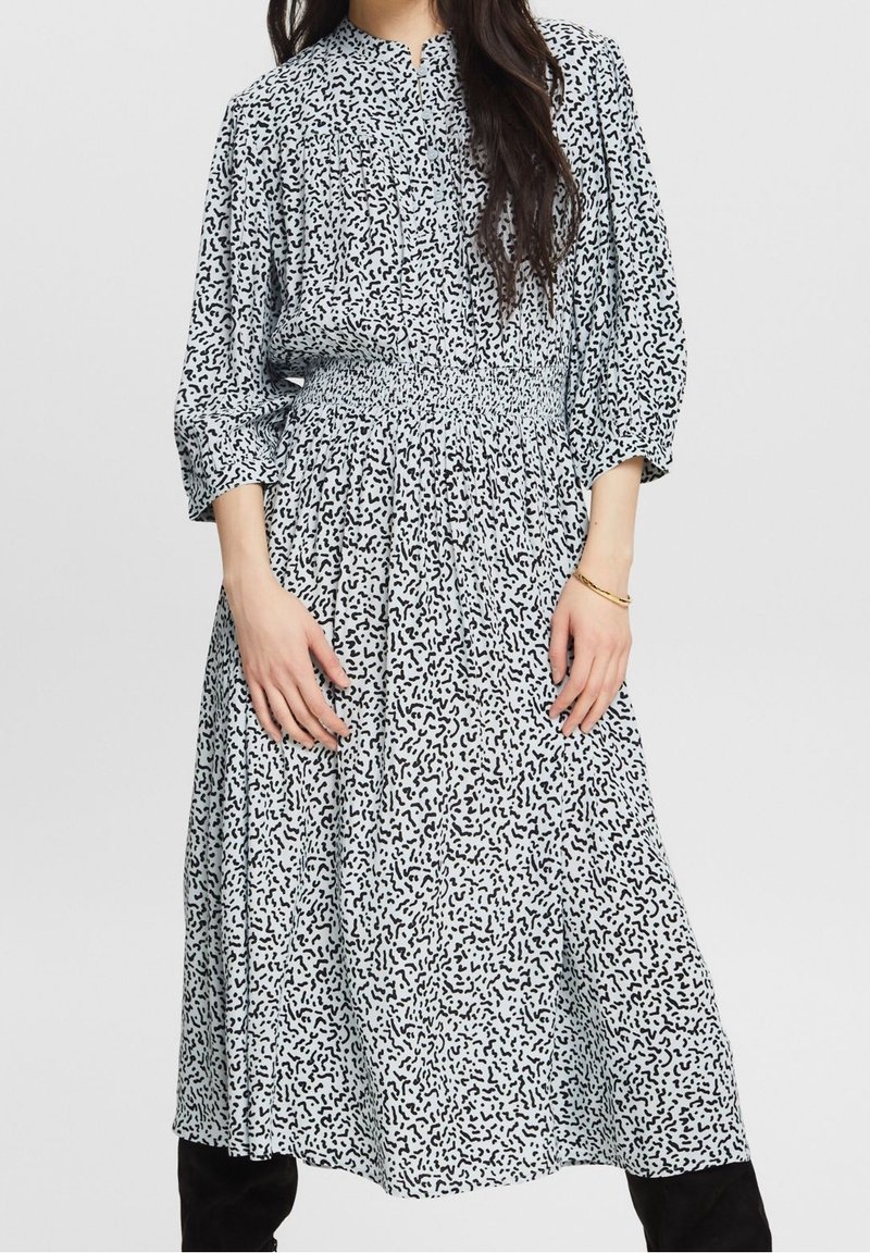 Femme portant une robe midi blanche avec un imprimé abstrait noir, manches 3/4 et taille smockée, associée à des bottes noires et un bracelet en or.