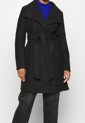 Mujer con un abrigo de lana gris oscuro ajustado con cinturón, sobre un suéter azul brillante y pantalones oscuros, posando frente a un fondo liso.