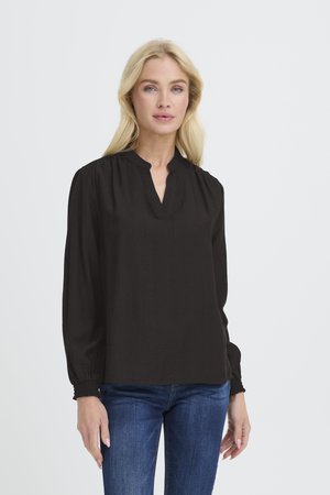 Blouse noire avec col montant, manches longues et texture froncée aux poignets. Tissu léger avec une finition lisse. Les jeans ne sont pas l'élément principal.