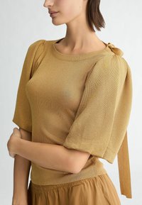 Maglione color senape con un morbido tessuto a maglia testurizzato, maniche a sbuffo, scollatura a costine e un dettaglio a fiocco sulla spalla, abbinato a una gonna marrone.