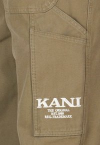 Poche cargo en tissu khaki avec le logo "KANI" en broderie blanche et un texte supplémentaire en dessous, mettant en avant les détails de matériau et de design.