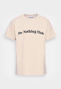 Béžové tričko s krátkým rukávem a černým textem "Do Nothing Club" zakřiveným písmem na hrudi a malou visačkou "On Vacation" uvnitř výstřihu.