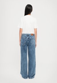 Femme aux longs cheveux noirs, debout de dos, portant un haut blanc à manches courtes et un jean bleu ample.