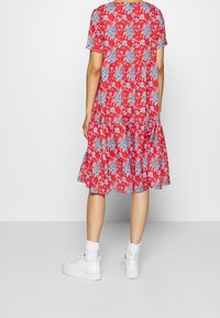 Robe fleurie rouge avec des manches courtes, bordure volantée, présentant des motifs floraux bleu et blanc. Portée avec des baskets blanches et des chaussettes de cheville.