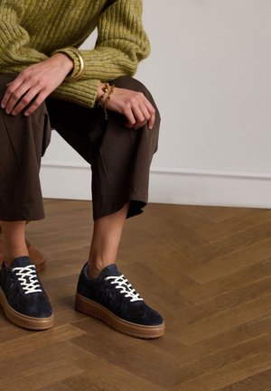 Marineblauwe suède sneakers met witte veters, voorzien van een stevige rubberen zool, gecombineerd met bruine wijde broek en een groene geribbelde trui.