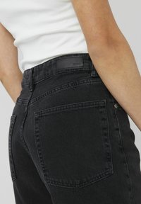 Jean en denim noir à taille haute, avec deux poches arrière et un patch en cuir au niveau de la taille. Fini texturé, coupe ajustée.