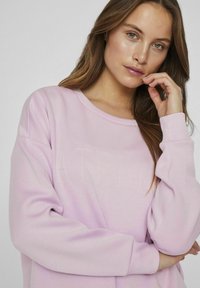 Sudadera de color rosa claro con corte relajado, que presenta letra bordada en un tono ligeramente más oscuro. Tejido suave y texturizado con cuello redondo.
