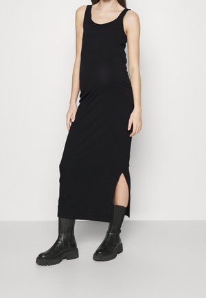 Schwangere Frau trägt ein ärmelloses schwarzes Midikleid mit seitlichem Schlitz und schwarzen klobigen Stiefeln, steht vor weißem Hintergrund.