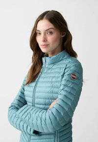 Helles blaues, gestepptes Jacket mit Reißverschluss und hohem Kragen. Verfügt über eine gepolsterte Textur und ein rundes Logobadge am linken Ärmel.