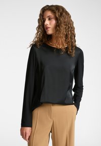 Schwarzes Langarmshirt aus glattem Stoff mit rundem Halsausschnitt. Kombiniert mit hellbeigen Hosen. Dezenter Glanz und lässige Passform.