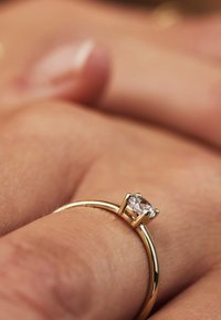 Goldring mit einem glatten, runden Band und einem rechteckigen, klaren Stein, der in vier Krappen gefasst ist, nah auf einem Finger abgebildet.