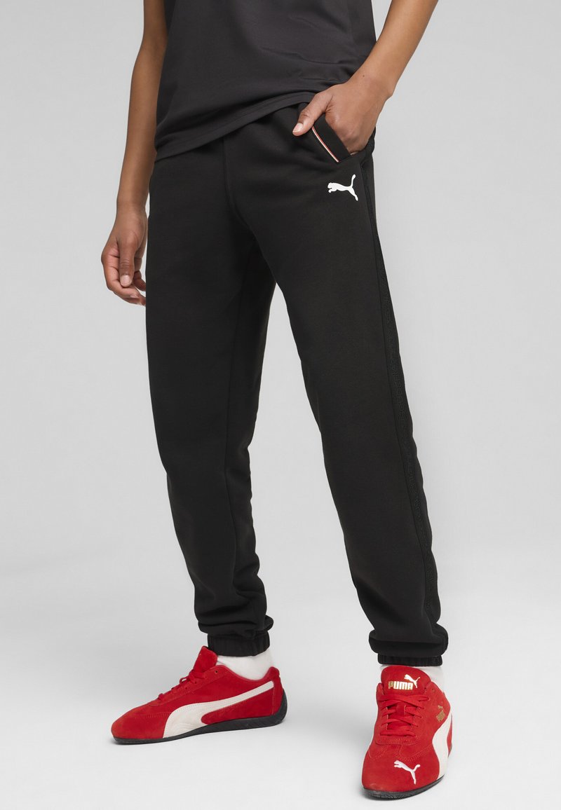 Pantalones de chándal negros con bolsillos laterales y puños elásticos, con un logo blanco de Puma. Combinados con zapatillas rojas y blancas de Puma.