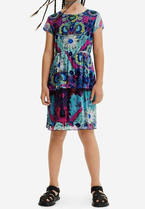 Enfant portant une robe à manches courtes, à motifs floraux et géométriques multicolores avec une jupe plissée et des sandales noires.