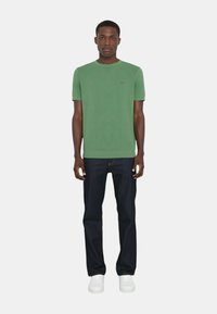 T-shirt verde a maniche corte con colletto rotondo e un piccolo logo, abbinato a jeans blu scuro e sneakers bianche, creando un look casual.