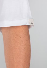 Cuff di camicia bianca a maniche corte con orlo arrotolato e etichetta in tessuto. L'etichetta presenta un logo con un design astratto. Texture di cotone morbido.