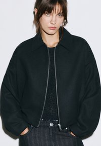 Geacă bomber neagră cu fermoar în față, croială oversized și țesătură texturată. Pur această geacă peste un pulover tricotat închis, cu guler.