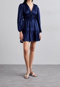 Námořnicky modré šaty s hlubokým výstřihem do V, nabíraným pasem a vícevrstvou sukní. Obsahuje dlouhé nabírané rukávy. Spojené se stříbrnými sandály na vysokém podpatku.