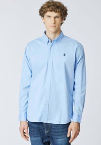 Giovane uomo con capelli ricci che indossa una camicia azzurra a maniche lunghe con bottoni e jeans scuri, in piedi davanti a uno sfondo bianco uniforme.