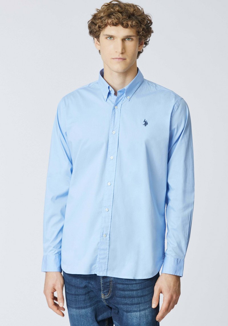 Giovane uomo con capelli ricci che indossa una camicia azzurra a maniche lunghe con bottoni e jeans scuri, in piedi davanti a uno sfondo bianco uniforme.