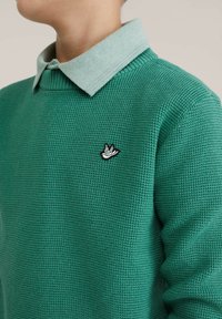 Pull vert tricoté avec un motif texturé, comprenant une chemise à col vert clair en dessous et un écusson brodé noir et blanc.