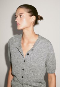 Cardigan grigio a maniche corte con chiusura a bottoni, tessuto a maglia testurizzato e un sottile motivo melangiato. Presenta un collo a V e una vestibilità rilassata.