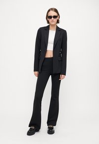Čierny šitý blazer, biele skrátené tričko, čierne rozšírené nohavice a masívne čierne mokasíny so zlatým retiazkou. Hladká, štruktúrovaná tkanina.