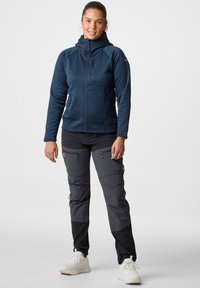 Marineblauwe zip-up jas met een capuchon, voorzien van ritssluitingen; gecombineerd met grijze broek met cargopockets en verstevigde onderbenen. Witte sneakers.