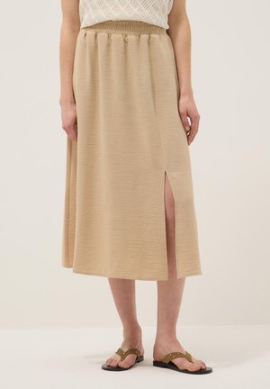 Jupe midi beige avec fente latérale, taille élastique, portée avec un haut en tricot blanc et des sandales plates marron avec des détails à œillets.