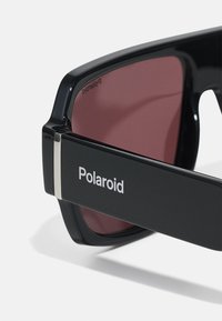 Ochelari de soare Polaroid negri cu rame dreptunghiulare, lentile roșii închise și un accent argintiu pe tâmplă cu logo-ul mărcii.