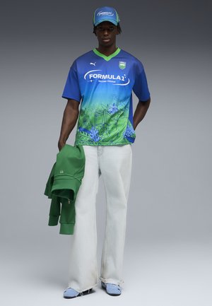 Person iført en blå og grøn gradient Formula 1 Racing League skjorte og kasket, hvide bukser med vide ben, lyseblå sko, holder en grøn jakke.