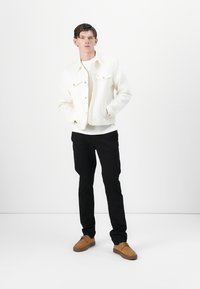 Hvit denimjakke over en kremfarget genser, kombinert med svarte bukser og brune sko. Modellen står med hendene i lommene mot en hvit bakgrunn.