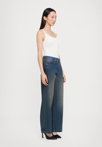 Široke modre denim hlače z bledim učinkom, v kombinaciji z oprijetim belim topom. Model nosi črne špicaste pete, stoječe v profilu.