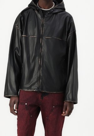 Persoon draagt een zwarte leren jas met capuchon en ritssluiting, en donkerrode, gestructureerde broek met steekzakken vooraan en klinknageldetails.